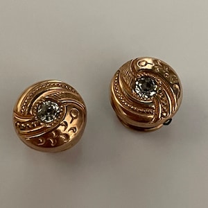 Könnte beinhalten: Ein Paar goldfarbene Manschettenknöpfe mit einem klaren Strassstein in der Mitte jedes Knopfes. Die Manschettenknöpfe haben ein wirbelndes Design.