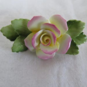 May include: A porcelain rose with yellow petals edged in pink, surrounded by green leaves. The flower is a detailed, handcrafted piece, likely for decorative purposes or jewelry.