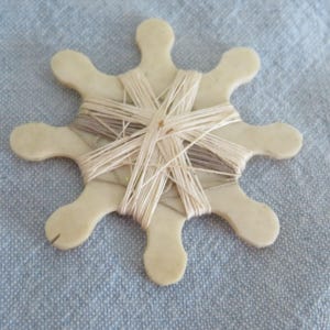 Può includere: Una bobina piatta a forma di stella color bianco sporco con otto braccia arrotondate, avvolta con filo da ricamo sottile color beige chiaro e marrone chiaro. La bobina ha una superficie liscia e opaca con una piccola tacca su un braccio.