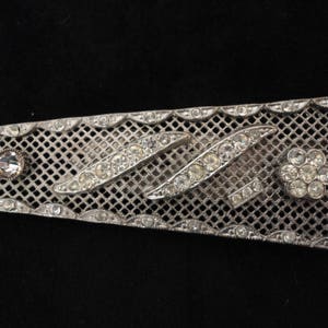 May include: A silver mesh brooch with a geometric design of rhinestones. The brooch has a flower-shaped rhinestone cluster in the center.