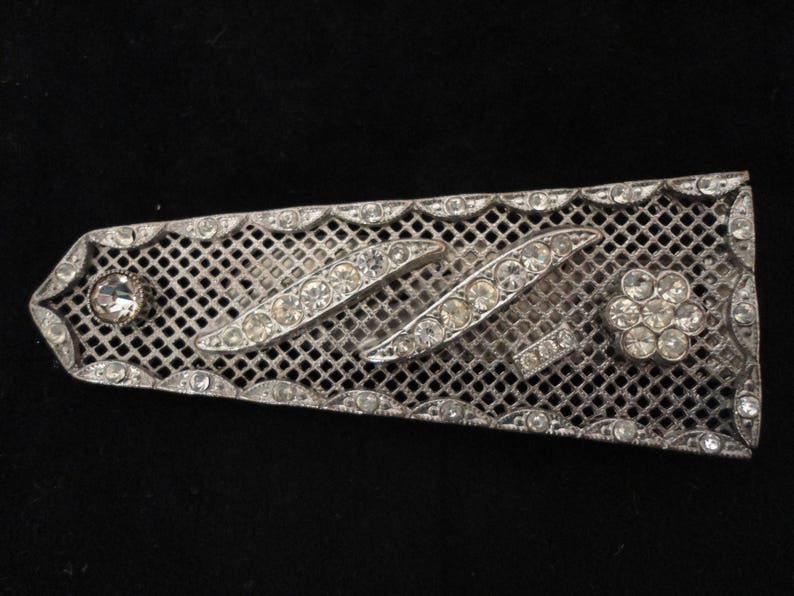 May include: A silver mesh brooch with a geometric design of rhinestones. The brooch has a flower-shaped rhinestone cluster in the center.