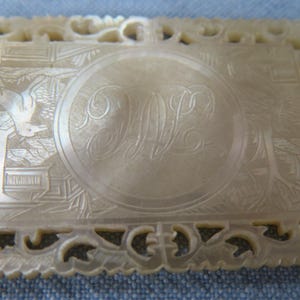 Peut inclure: Plaque rectangulaire blanc cassé avec une bordure finement sculptée. Le centre présente un ovale avec des initiales gravées. Des sculptures détaillées incluent un oiseau, des bâtiments et du feuillage.