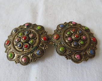 May include: A pair of ornate gold-toned metal belt buckles with a circular design. The buckles are decorated with a variety of colorful enamel dots in shades of red, green, blue, and pink.