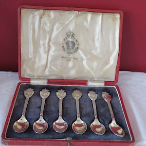 Pode incluir: Um conjunto de seis colheres de chá de prata em uma caixa forrada de veludo vermelho. As colheres são decoradas com um brasão real e a inscrição "Coronation Day 1937".