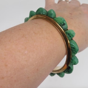 May include: A gold-tone bangle bracelet with a series of green, rounded, textured stones. The bracelet is worn on a person's arm, showcasing the contrast between the gold and green. The stones are a vibrant shade of green.