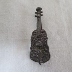May include: A detailed, antique-style violin-shaped brooch. The brooch is made of a dark, possibly tarnished metal, with intricate floral and scrollwork details. The design includes the violin's body, neck, and scroll.