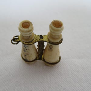 Puede incluir: Miniatura de anteojos de ópera de color marfil con un marco de metal color latón. Los pequeños binoculares tienen un diseño detallado y probablemente sean antiguos. Las lentes parecen ser de color ámbar claro. El artículo está sobre un fondo blanco.