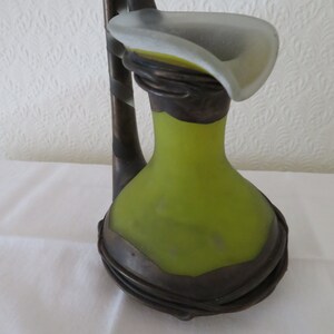 May include: A green glass vase with a brown metal handle and base. The vase has a unique shape and a frosted glass top.