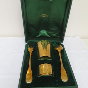 Könnte beinhalten: Ein Vintage-Besteck-Set in Goldoptik, einschließlich Gabel, Löffel, Serviettenring und Becher, präsentiert in einem grün samtgefütterten Etui. Das Etui ist dunkelgrün mit goldfarbener Hardware. Das Besteck hat ein Muscheldesign an den Griffen.