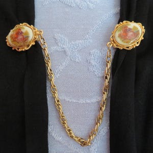 May include: Vintage gold-tone sweater clips. Each oval clip has a painted scene in orange, brown, and cream, framed by gold filigree. A gold chain connects the two clips. The clips are designed to fasten clothing.