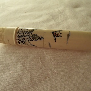 Puede incluir: Objeto cilíndrico de color marfil con un extremo texturizado. El cuerpo está decorado con ilustraciones en blanco y negro de una abeja y una rama con elementos circulares. El objeto está sobre una superficie blanca.