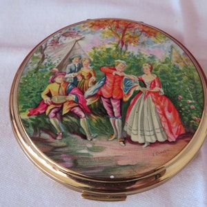 May include: A round, gold-toned compact with a painted scene of five people in a forest setting. The scene features a man playing a lute, two women, and two men. The compact has a white background with a gold rim.