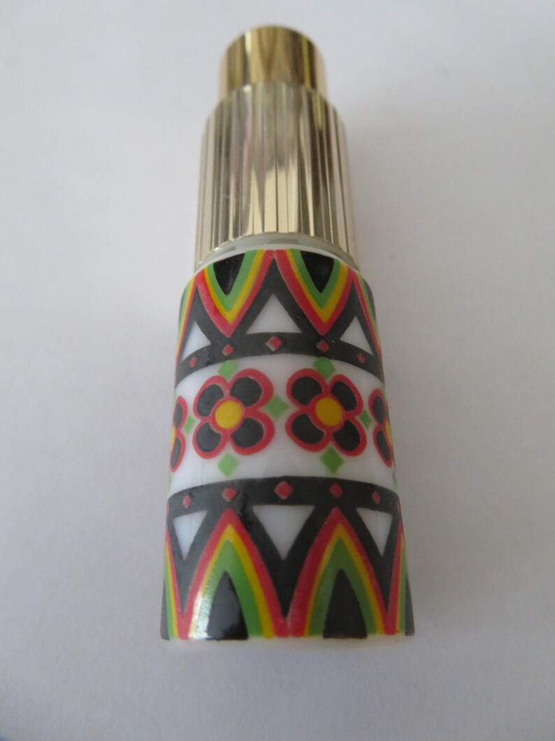 May include: A decorative perfume bottle with a gold-colored cap and a patterned body. The body features a white base with black, red, yellow, and green geometric designs and floral accents. The bottle is approximately 4 inches tall.