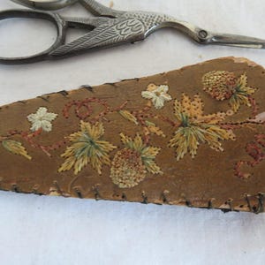 May include: A brown leather needle case with embroidered floral designs in shades of green, yellow, and red. The case is slightly damaged and has a frayed edge.