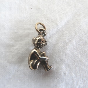 Könnte beinhalten: Ein kleiner, goldfarbener Anhänger in Form einer sitzenden Figur. Der Anhänger hat oben eine Schlaufe zum Befestigen an einer Kette oder einem Armband. Die Figur ist mit Gesichtszügen detailliert und hält ihre Knie.