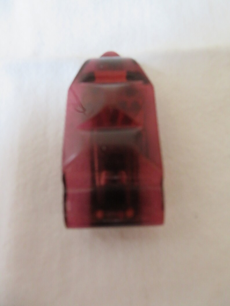 May include: A small, translucent, red plastic toy car. The car has a rounded shape and a slightly pointed front end.