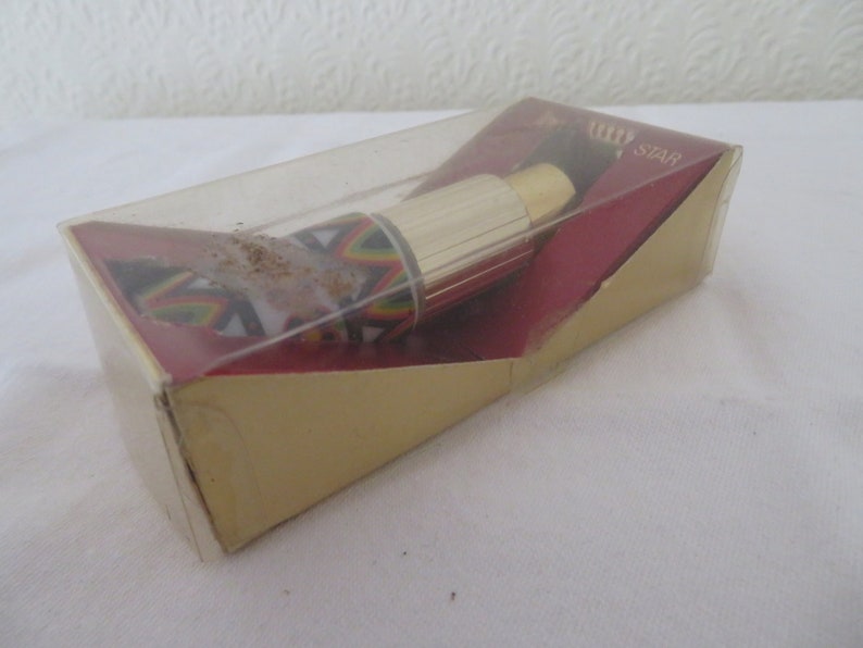 May include: A vintage lipstick in a clear plastic case, inside a gold and red box. The lipstick has a gold metal tube and a colorful geometric pattern on the base. The box has the word "STAR" printed on it.