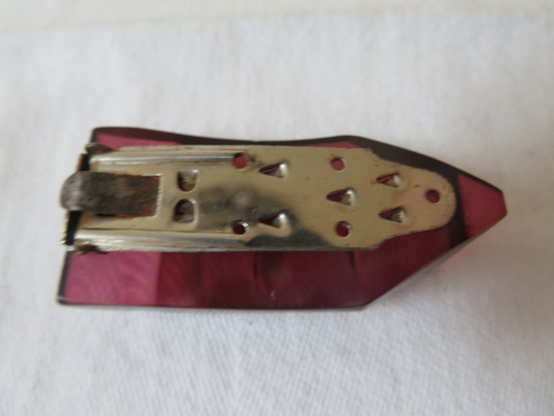 May include: A vintage purple glass brooch with a metal clasp. The clasp has a decorative design with small holes and a textured surface.