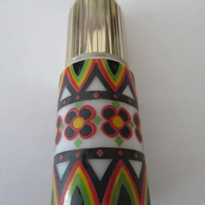 May include: A decorative perfume bottle with a gold-colored cap and a patterned body. The body features a white band with black, red, yellow, and green geometric designs and floral patterns. The bottle is cylindrical and stands upright.