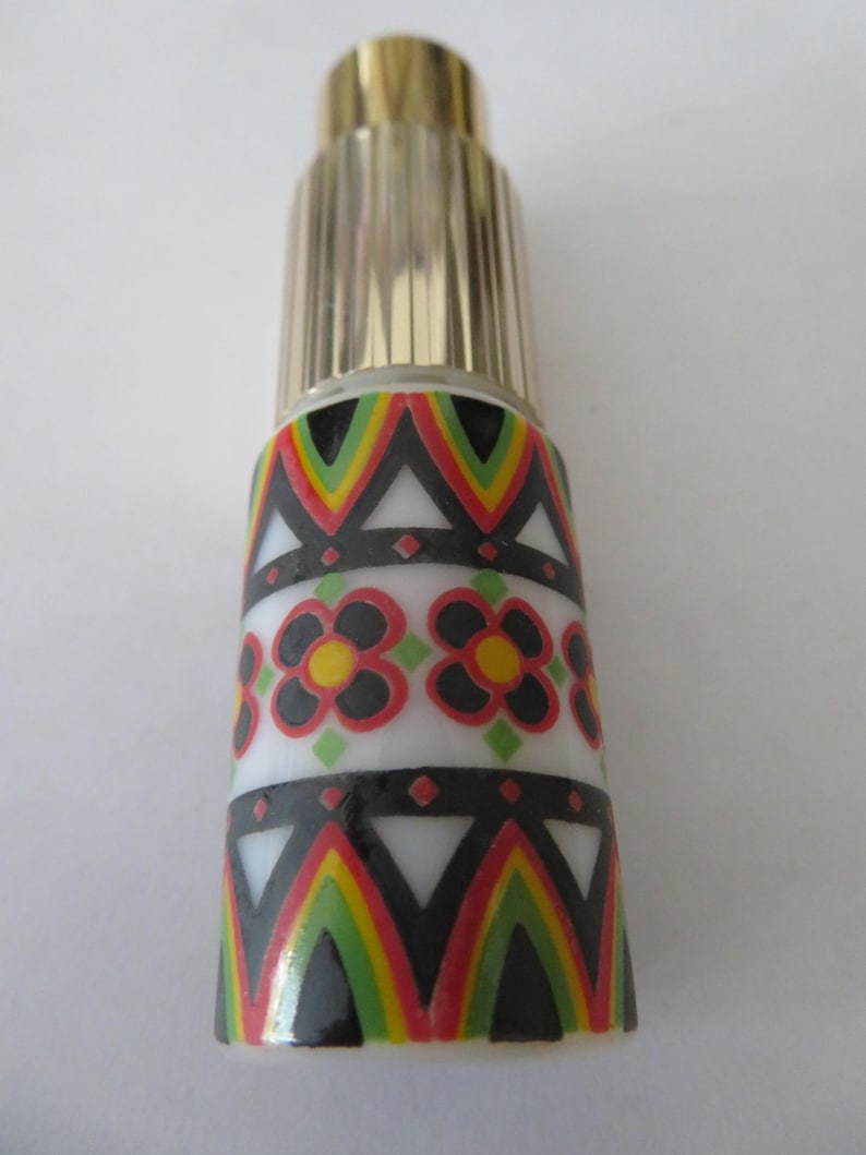 May include: A decorative perfume bottle with a gold-colored cap and a patterned body. The body features a white band with black, red, yellow, and green geometric designs and floral patterns. The bottle is cylindrical and stands upright.