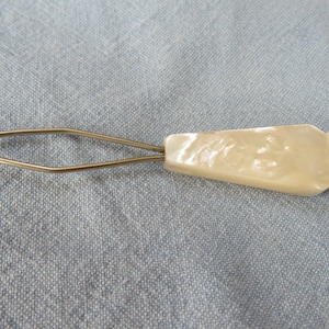 Puede incluir: Un gancho para botones vintage con un mango nacarado y un lazo de alambre. El mango es de color crema claro y tiene una superficie ligeramente texturizada. El lazo de alambre es de color dorado.