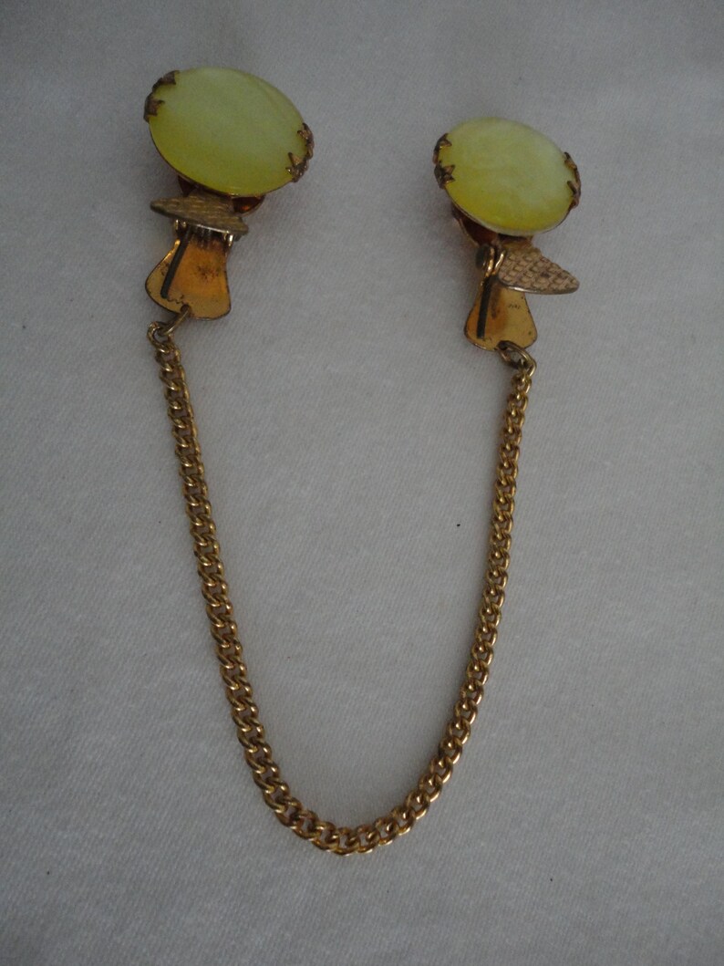 May include: A pair of gold tone clip-on earrings with a pale yellow glass stone. The earrings are connected by a gold chain.