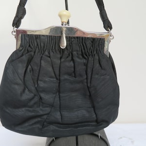 以下が含まれることがあります： 銀色の留め具と白いプラスチック製のトップハンドルが付いた、黒いサテンのイブニングバッグです。