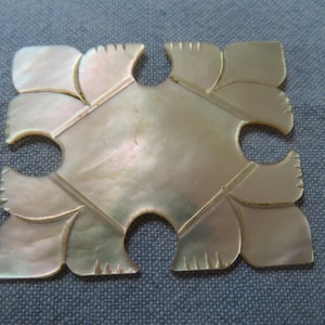 May include: A mother-of-pearl puzzle piece with a unique design. The piece has a central square shape with curved edges and leaf-like extensions. The iridescent surface reflects light, creating a shimmering effect. The piece is set against a blue background.