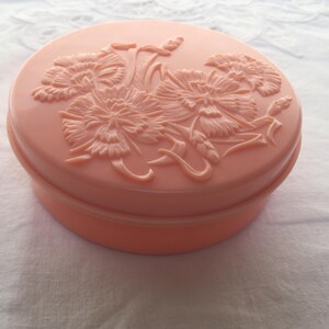 Può includere: Un portagioie rotondo color pesca con un motivo floreale sul coperchio. Il coperchio presenta un rilievo in rilievo di fiori e foglie. La scatola è su una superficie bianca strutturata.