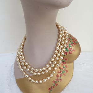 May include: Three-strand pearl necklace displayed on a mannequin bust. The bust is painted gold with a floral design and a neutral-toned face. The pearls are a creamy off-white color, creating a classic and elegant look.