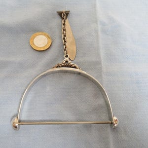 May include: An antique silver-coloured garment hanger with a decorative design. The hanger features a curved bar with a horizontal rod and a chain attached to a decorative top. A coin is next to the hanger.