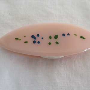 Pode incluir: Uma lançadeira de frivolité ovalada rosa pálido com um padrão decorativo de pontos azuis e verdes. Um fio branco é visível, estendendo-se da lançadeira.