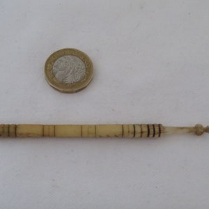 Puede incluir: Un primer plano de una pequeña aguja de tejer de color marfil con marcas negras. La aguja es aproximadamente del tamaño de una moneda británica de una libra.