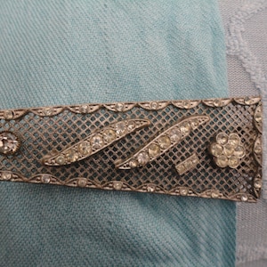May include: A silver-toned brooch with a geometric design featuring a mesh pattern and clear rhinestones. The brooch has a flower-shaped design with rhinestones in the center.