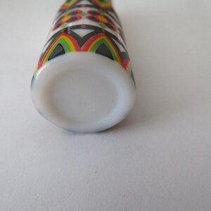 May include: A white bottle with a colorful geometric pattern in black, red, yellow, and green. The bottle has a rounded base and a cylindrical shape. The design is repeated around the bottle.