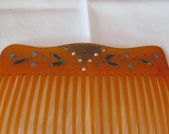 May include: An orange plastic comb with a decorative floral design and silver and white accents. The comb has a curved top and a row of teeth.