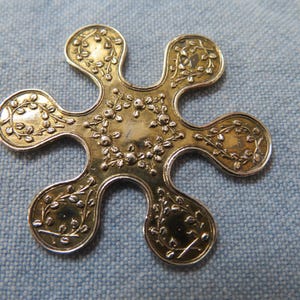 May include: A gold-toned metal decorative piece with a six-petal design. Each petal features an embossed floral pattern with leaves and small flowers. The center of the piece also has a floral design. The piece is set against a blue fabric background.
