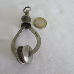 May include: A silver-toned metal caliper tool with a threaded adjustment screw at the top and a circular ring for hanging. The tool features two curved arms and two rounded contact points at the base, positioned next to a gold-coloured coin for scale.