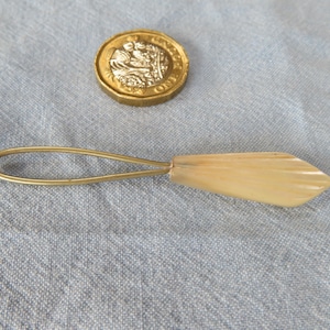 Puede incluir: Un pequeño cuchillo de mantequilla vintage con un mango en forma de hoja de color claro y un lazo de metal dorado. Una moneda de oro está al lado del cuchillo. El cuchillo está sobre una tela azul claro.