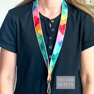 Peut inclure: Un cordon tour de cou arborant un motif géométrique multicolore vif avec un dégradé arc-en-ciel de triangles et de petites croix blanches. La sangle en tissu est fixée à un mousqueton pivotant en métal argenté. Le texte TEN COW QUILTS est visible.