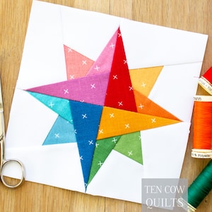 Può includere: Un blocco di quilt a forma di stella multicolore su sfondo bianco. La stella è composta da pezzi di tessuto rosa, rosso, arancione, giallo, verde, blu e viola. Forbici e rocchetti di filo su una superficie di legno.