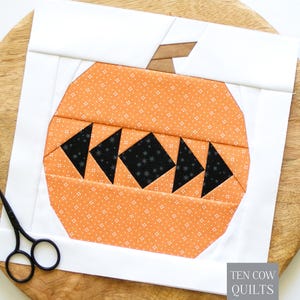May include: A square fabric quilt block featuring an orange pumpkin design with a white background. The pumpkin has a brown stem and a black face made of geometric shapes. Black scissors are positioned on the left. Text reads TEN COW QUILTS.