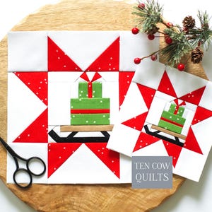 May include: Two square fabric quilt blocks featuring a red and white star pattern surrounding a central green gift box on a brown sled. The blocks are white, red, and green with small white speckles. A pair of black scissors is visible.