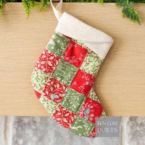 May include: A quilted Christmas stocking with a patchwork design. The stocking features red and green squares with floral and holiday-themed patterns. It has a cream-colored cuff and hanging loop. The text "TEN COW QUILTS" is visible.