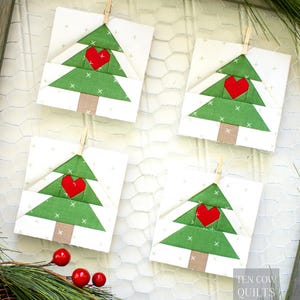 Könnte beinhalten: Vier quadratische Stoffornamente mit einem grünen Weihnachtsbaumdesign und einem roten Herz in der Mitte. Die Bäume sind mit einer Holzklammer auf einem weißen Hintergrund befestigt. Der Hintergrund ist weißer Maschendraht.