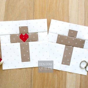 May include: Two white and tan fabric quilt blocks with cross designs. One block features a red heart in the centre of the cross. The blocks are on a wooden surface. The text "TEN COW QUILTS" is visible.