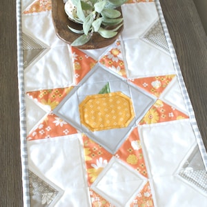 Könnte beinhalten: Ein weißer Tischläufer mit Patchwork-Design aus orangefarbenem, gelbem und grauem Stoff. In der Mitte des Läufers befindet sich eine graue und gelbe Kürbis-Applikation. Der Läufer hat einen grauen und weißen Gingham-Besatz.