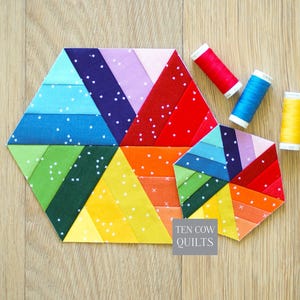 May include: Two hexagonal fabric quilt blocks featuring a rainbow color gradient with white speckled patterns, accompanied by three spools of red, blue, and yellow thread. A gray square label displays the text TEN COW QUILTS in white.
