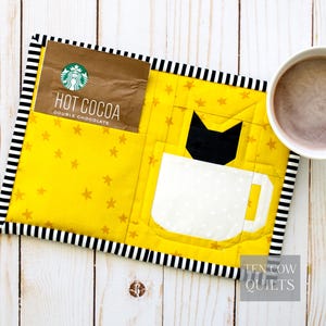 Könnte beinhalten: Ein gelber, gesteppter Becher-Untersetzer mit einem schwarz-weiß gestreiften Rand. Der Untersetzer zeigt eine weiße Tasse mit einer schwarzen Katze und eine Tasche mit einem Starbucks-Kakao-Päckchen. Rechts steht eine Tasse heiße Schokolade.