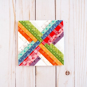 K&ouml;nnte beinhalten: Ein farbenfroher Patchwork-Quiltblock mit einem Kreuzmuster. Der Block besteht aus verschiedenen Stoffen in Rot-, Orange-, Gelb-, Gr&uuml;n-, Blau- und Violett&ouml;nen.